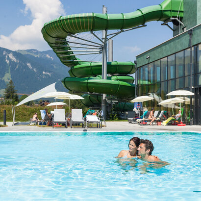 Das Bild erfasst eine lebhafte Szene in einem Resort. Der Schwerpunkt liegt auf einer großen, grünen Wasserrutsche, die gerade in Gebrauch ist. Die Rutsche ist gekrümmt und erscheint aus einem robusten Material gefertigt zu sein. Sie befindet sich in der Mitte des Bildes und zieht die Aufmerksamkeit auf sich.  Im Vordergrund schwimmen zwei Personen in einem Becken. Sie stehen einander nahe, was darauf hindeutet, dass sie vielleicht gemeinsam das Wasserrutschenvergnügen genießen. Das Becken selbst ist ein lebhaftes Blau und kontrastiert gut mit dem Grün der Rutsche.  Der Hintergrund des Bildes offenbart eine schöne Berglandschaft. Die Berge sind grün und üppig, was der Resortszenerie einen Hauch von Natur verleiht.  Das Resort selbst wird durch einen großen Gebäudekomplex mit einem grünen Dach gekennzeichnet. Dieses Gebäude ist wahrscheinlich Teil der Resorteinrichtungen und beherbergt Amenities wie ein Restaurant oder ein Geschäft.  Insgesamt zeichnet das Bild ein Bild von einem unterhaltsamen und erholsamen Tag in einem Resort, wo Menschen die Wasserrutsche genießen und die schöne Bergkulisse erfreuen.