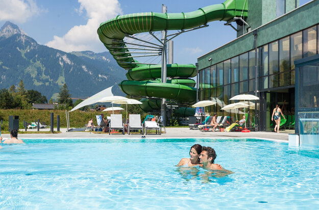 Das Bild erfasst eine lebhafte Szene in einem Resort. Der Schwerpunkt liegt auf einer großen, grünen Wasserrutsche, die gerade in Gebrauch ist. Die Rutsche ist gekrümmt und erscheint aus einem robusten Material gefertigt zu sein. Sie befindet sich in der Mitte des Bildes und zieht die Aufmerksamkeit auf sich.  Im Vordergrund schwimmen zwei Personen in einem Becken. Sie stehen einander nahe, was darauf hindeutet, dass sie vielleicht gemeinsam das Wasserrutschenvergnügen genießen. Das Becken selbst ist ein lebhaftes Blau und kontrastiert gut mit dem Grün der Rutsche.  Der Hintergrund des Bildes offenbart eine schöne Berglandschaft. Die Berge sind grün und üppig, was der Resortszenerie einen Hauch von Natur verleiht.  Das Resort selbst wird durch einen großen Gebäudekomplex mit einem grünen Dach gekennzeichnet. Dieses Gebäude ist wahrscheinlich Teil der Resorteinrichtungen und beherbergt Amenities wie ein Restaurant oder ein Geschäft.  Insgesamt zeichnet das Bild ein Bild von einem unterhaltsamen und erholsamen Tag in einem Resort, wo Menschen die Wasserrutsche genießen und die schöne Bergkulisse erfreuen.