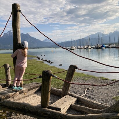 Das Bild fängt eine friedliche Szene in einer Marina ein. Ein junges Mädchen steht auf einem hölzernen Boot. Sie ist in einem pinkfarbenen Oberteil und pinkfarbenen Hosen gekleidet. Das Bild wird durch eine Reihe anderer Boote im Hintergrund, die an der Marina liegen, ergänzt, vor dem atemberaubenden Panorama einer Berglandschaft. Das klare Blau des Himmels über ihr unterstreicht die allgemeine Ruhe der Szene. Die Position des Mädchens auf dem Boot und ihre feste Umklammerung des hölzernen Pols lassen darauf schließen, dass sie ihren Aufenthalt auf dem Wasser genießt. Das Bild fängt einen Moment der Erholung und Entdeckung für das junge Mädchen wunderschön ein.