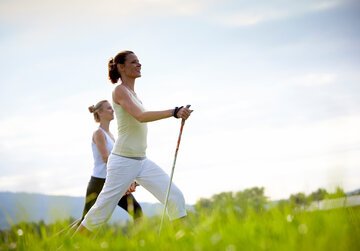 Das Bild zeigt zwei sportlich gekleidete, schlanke Frauen, die mit Nordic-Walking-Stöcken über eine grüne Wiese laufen. Beide Frauen haben die Haare zusammengebunden und blicken glücklich voraus. Der blaue Himmel ist von sanften Wolken durchzogen.