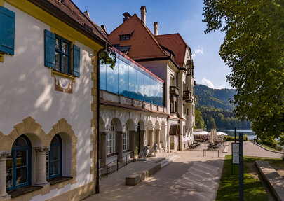 Man blickt entlang des Gebäudes des Museums der bayerischen Könige. Im Hintergrund erheben sich die Berge. Der Bergwald beginnt, davor ist der Alpsee zu sehen. Am Ufer des Alpsees sieht man die Terrasse des Restaurants Alpenrose am See. Unter Sonnenschirmen sieht man mehrere Personen an Tischen sitzen. Links und rechts neben dem grünen Portal des Museums stehen zwei lebensgroße, steinerne Löwen.  