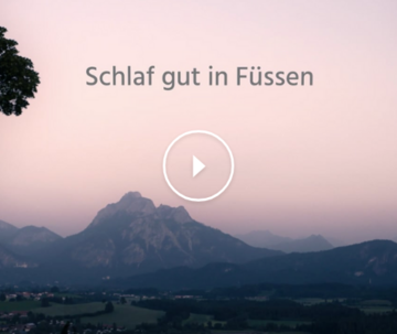 Das Bild zeigt eine in Abendlicht getauchte Landschaft mit einer imposanten Bergkulisse im Hintergrund. Der Himmel leuchtet in Lila- und Blautönen des letzten Sonnenlichts.