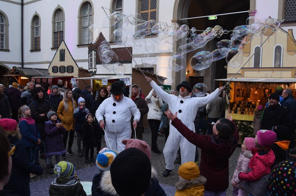 Das Bild zeigte zwei Männer, die als Schneemänner verkleidet sind. Die haben lange Holzstäbe in den Händen und lassen große Seifenblasen entstehen. Um sie herum stehen Kinder und Erwachsene, die sich die Darbietung ansehen. Im Hintergrund stehen Verkaufsbuden eines Adventsmarkts, die vor einem Gebäude platziert wurden. Es ist hell.  