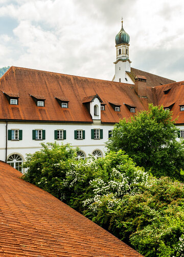 Das Bild zeigt das Franziskanerkloster in Füssen mit seinem markanten roten Ziegeldach und weißen Fassade. Üppige Bäume umgeben das Kloster.