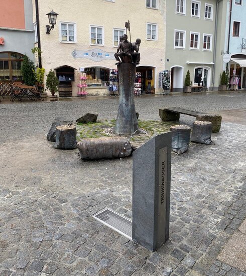 Das Bild zeigt eine Stadtstraßenszene. Im Zentrum des Bildes steht eine große, graue Statue. Die Statue besteht aus einer grauen Säule auf deren oberen Ende drei Männer sitzen. In die Säule ist die Form von Getreideähren eingearbeitet. Die Statue ist von einer Reihe kleiner Bänke umgeben. Einige der Bänke haben die Form von Mehlsäcken. Die Statue wird zum zentralen Punkt des Bildes, sie lässt unsere Aufmerksamkeit auf ihre Position richten. Es handelt sich hier um den Bäckerbrunnen auf dem Füssener Schrannenplatz. Im Vordergrund des Bildes ist eine schmale, graue und niedrige Säule zu sehen, auf der das Wort „Trinkbrunnen“ zu sehen ist. Aus dem Trinkbrunnen sprudelt ein kleiner Wasserstrahl nach oben.  Im Hintergrund sind Gebäude verschiedener Farben zu sehen, wie gelb, blau und weiß. Die Gebäude scheinen gewerblich zu sein. Man kann Schaufenstern erkennen. Die Straße selbst ist mit Kopfsteinpflaster gepflastert, was der Szene eine historische und charmante Atmosphäre verleiht. Der Himmel ist bedeckt, was darauf schließen lässt, dass das Wetter kühl oder bewölkt sein könnte. 