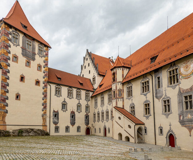 Das Bild zeigt den Innenhof des Hohen Schlosses in Füssen, der durch seine Illusionsmalerei an den Fassaden beeindruckt. Das Schlossgebäude hat rote Türen und ein Ziegeldach.