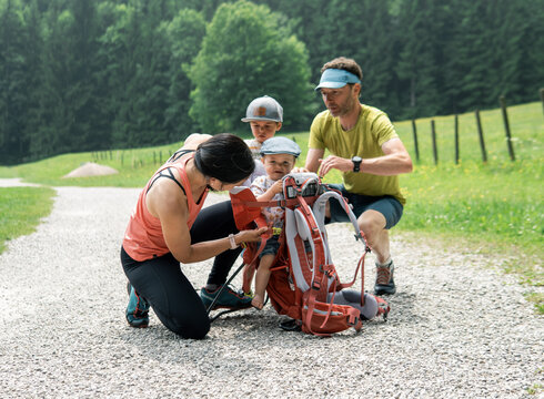 Eine Frau befestigt ein Kleinkind in einem Wanderrucksack, während ein Mann und ein weiteres Kind zuschauen. Die Szene spielt sich auf einem Kiesweg in einer grünen Landschaft ab, umgeben von Bäumen und Wiesen.