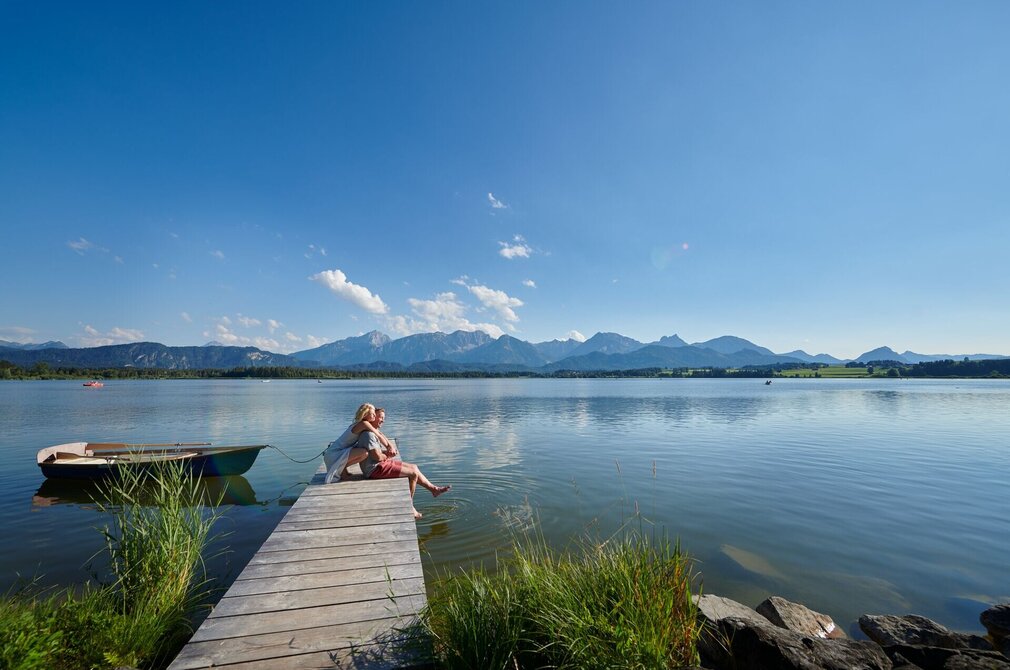 Auf dem Bild ist eine idyllische Szene am Hopfensee zu sehen. Es ist Sommer. Eine Frau kniet hinter einem Mann auf einem Holzsteg und umarmt ihn von hinten. Er sitzt auf dem Holzsteg. Seine Beine baumeln über dem Rand des Stegs herunter. Seine Füße berühren leicht die Wasseroberfläche des tiefblauen Sees. Beide blicken in die Ferne und lächeln. Der Holzsteg ragt in das ruhige Wasser des Hopfensees hinein, welcher von Bergen umgeben ist. Der Himmel ist blau und nur leicht durchzogen von wenigen Wolken. In der Ferne sind ein paar Ruder- und Tretboote auf dem Wasser zu sehen.