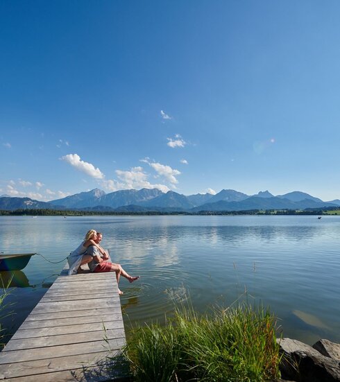 Auf dem Bild ist eine idyllische Szene am Hopfensee zu sehen. Es ist Sommer. Eine Frau kniet hinter einem Mann auf einem Holzsteg und umarmt ihn von hinten. Er sitzt auf dem Holzsteg. Seine Beine baumeln über dem Rand des Stegs herunter. Seine Füße berühren leicht die Wasseroberfläche des tiefblauen Sees. Beide blicken in die Ferne und lächeln. Der Holzsteg ragt in das ruhige Wasser des Hopfensees hinein, welcher von Bergen umgeben ist. Der Himmel ist blau und nur leicht durchzogen von wenigen Wolken. In der Ferne sind ein paar Ruder- und Tretboote auf dem Wasser zu sehen.