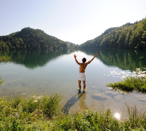 Das Bild zeigt einen Mann, der sich nur ein weißes Handtuch um die Hüften gebunden hat. Er steht im knöcheltiefen Wasser eines Sees und streckt die Arme nach oben.