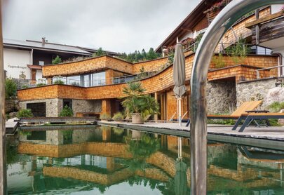 Das Bild zeigt ein Haus, dessen Fassade teilweise mit Schindeln, teilweise mit groben Steinen verkleidet ist. Davor liegt ein Pool. Das Haus hat mehrere Gebäudeteile, Terrasse und Balkone. Es wirkt wie eine ausgewogene Mischung aus Moderne und Tradition.