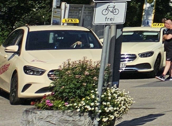 Das Bild zeigt das Gebäude des Füssener Bahnhofs. Links neben dem Gebäude erstreckt sich der Busbahnhof. Im Vordergrund stehen einige Taxen. Der Himmel ist blau. Die Sonne scheint. 